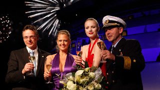 Taufe AIDAluna Franziska Knuppe hält einen Blumenstrauß und ein Sektglas in der Hand, im Hintergrund ist Feuerwerk zu sehen
